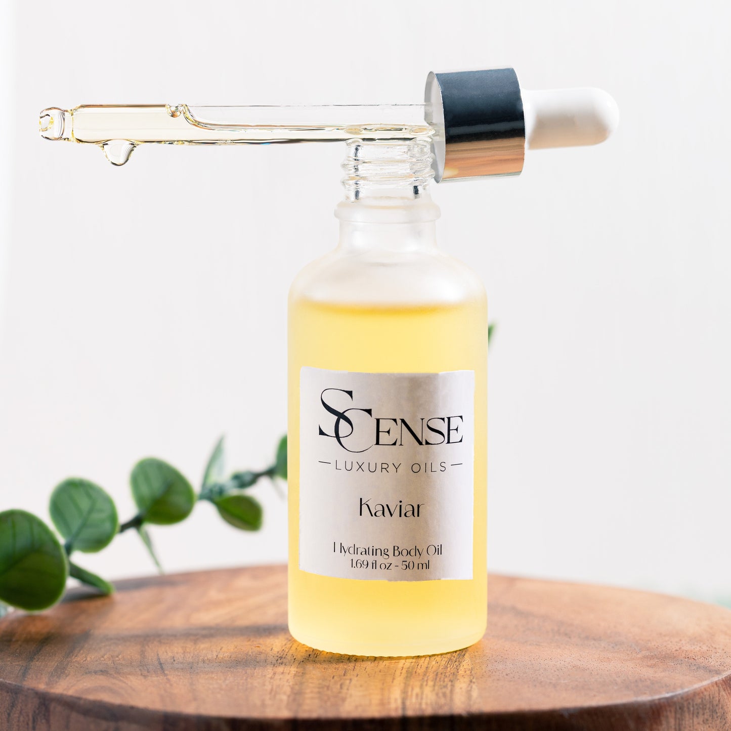 SCense Kaviar body oil in dropper bottle with the cap balancing on the top showing a golden oil inside the dropper, the bottle is placed on a wooden tray with a green leaf in soft natural light