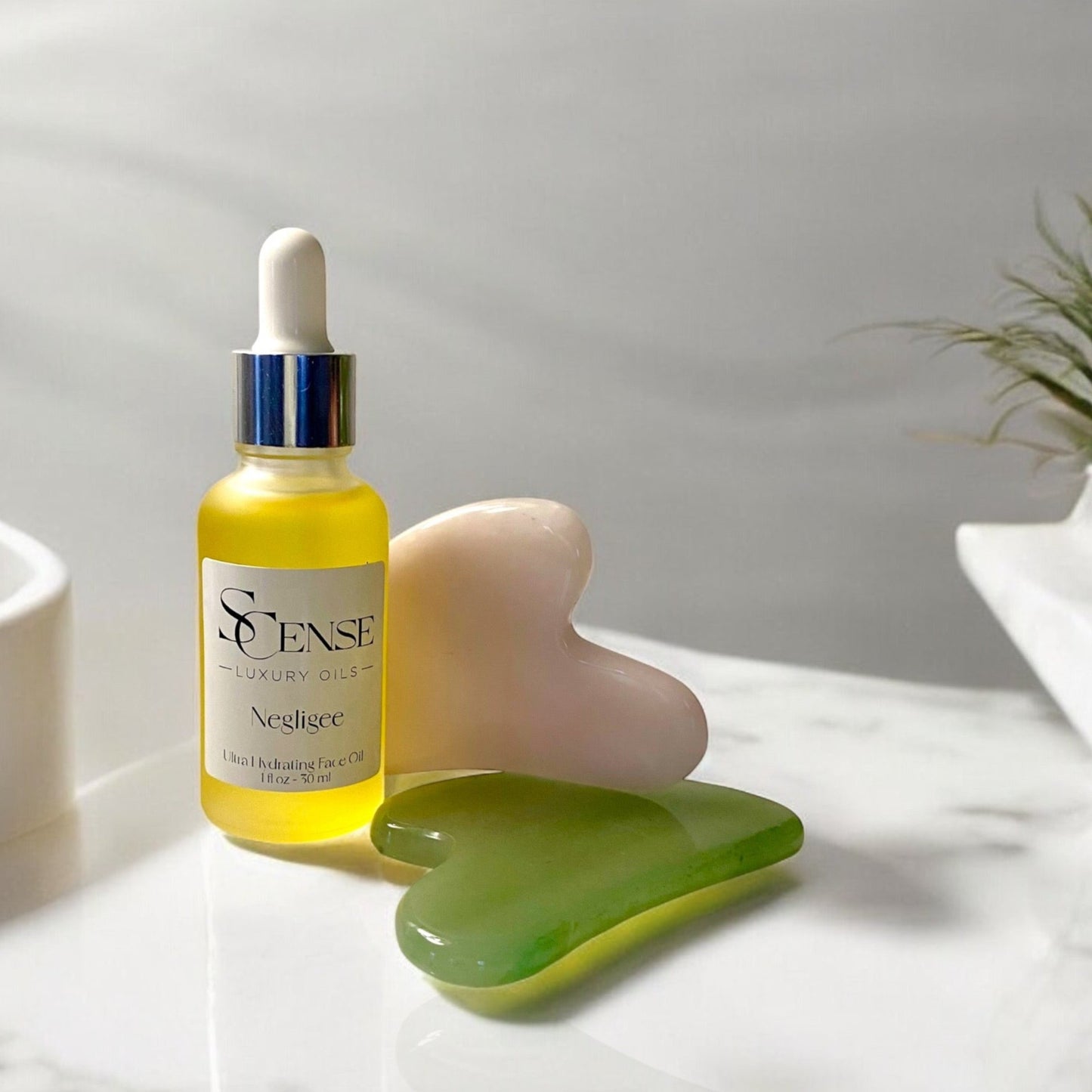 SCense Negligee oil in a frosted dropper bottle beside a pink quartz and green jade gua-sha facial massaging tools on a white counter top in natural light