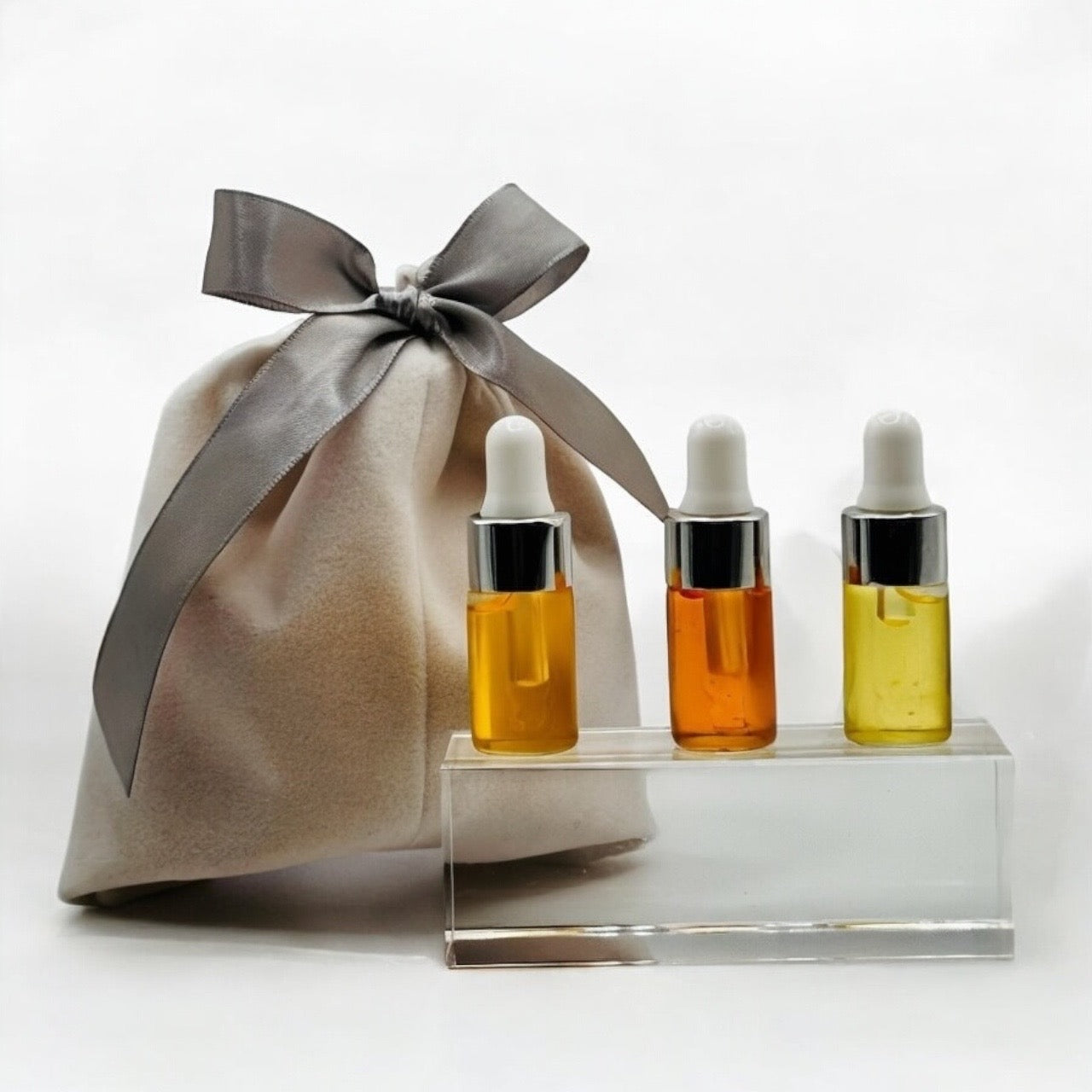 3 mini dropper bottles 3ml each filled with our signature facial oils set on a clear block in front of a soft white velvet pouch with silver ribbon