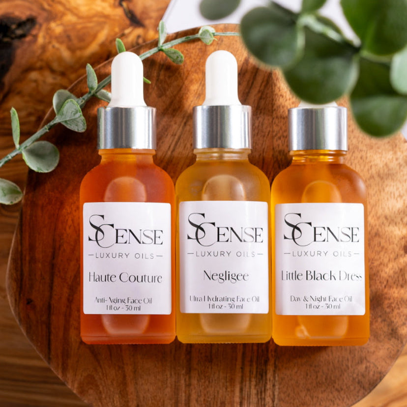 3 dropper bottles filled with Haute Couture, Negligee and Little Black Dress facial oils, flat lay on a round wooden board, surrounded by soft green eucalyptus stem 